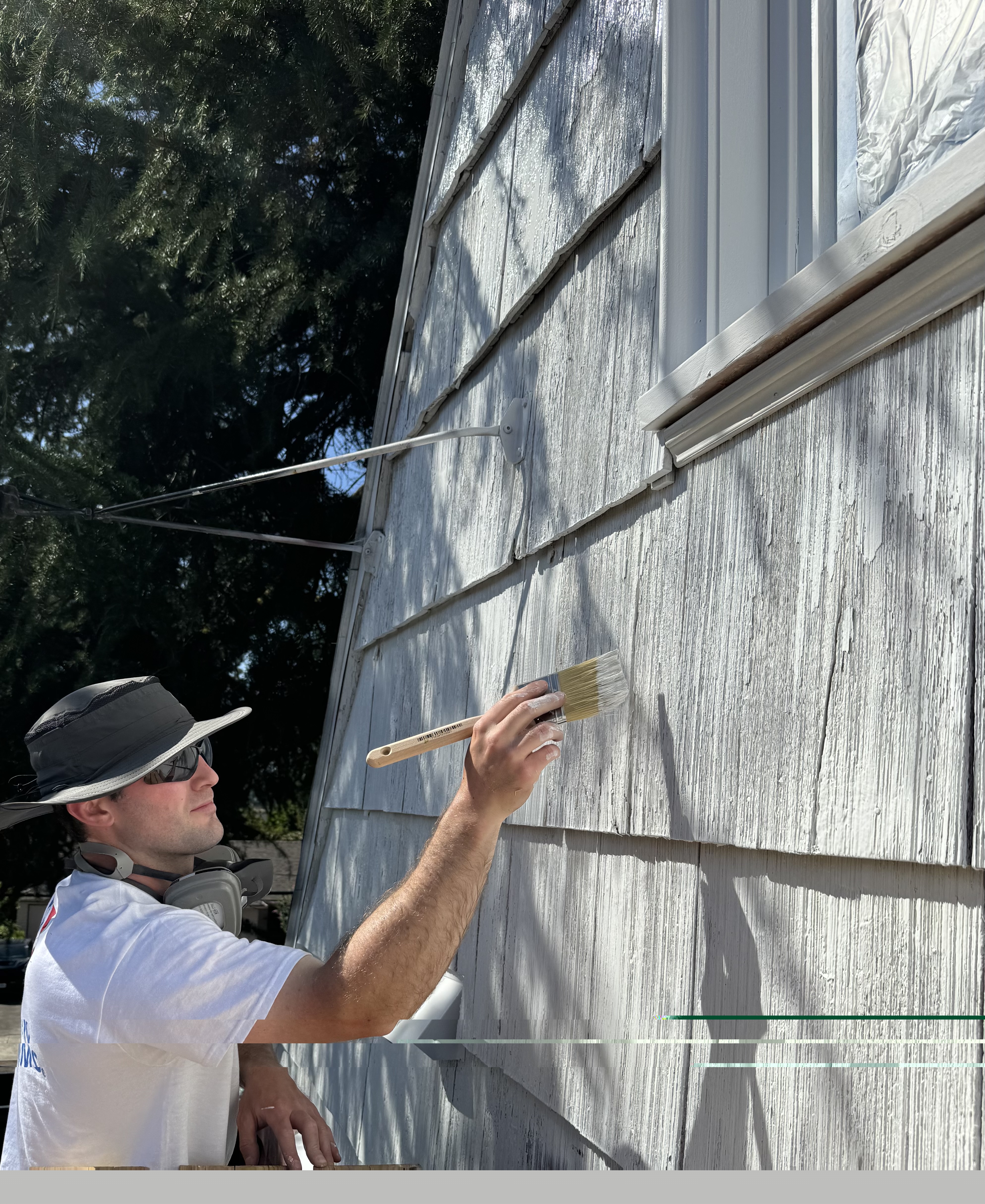 Man wearing a hat and sunglasses applies white paint to grey wooden house shingles.